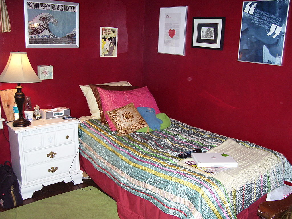 The Online Photo Dictionary: Teen girl's bedroom - wall, poster, curtain, piece of wood, bottle of water, lamp, night stand, drawers, clock radio, cup, vase, cushions, box, school bag, floor, rug, bed, blanket, picture, picture frame, laptop computer, remote control, cell phone, magazine